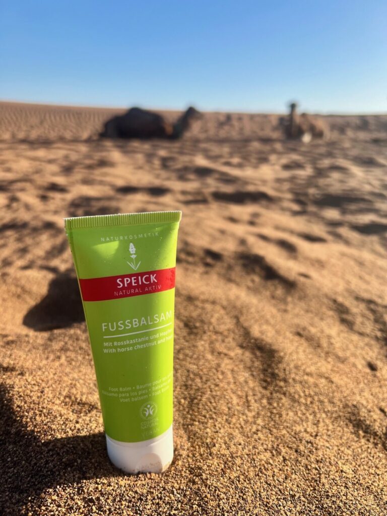 Speick Natural Aktiv Fußbalsam im Wüstensand, im Hintergrund liegende Dromedare